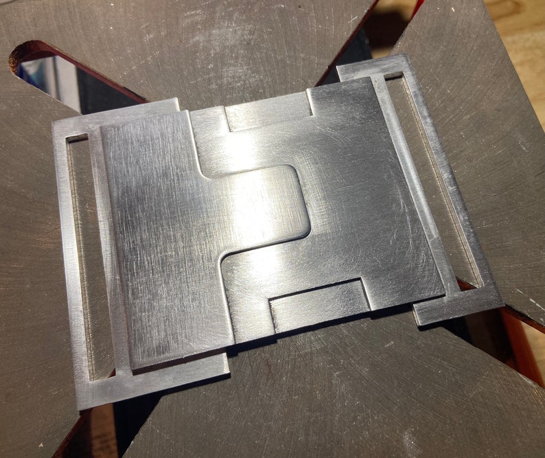 K&ouml;nnte beinhalten: Eine silberne Metallschnalle mit einem einzigartigen, ineinandergreifenden Design. Die Schnalle hat eine geb&uuml;rstete Oberfl&auml;che und ist aus hochwertigem Metall gefertigt.