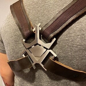 May include: A brown leather harness with a silver metal spider-shaped buckle. The harness has two straps that cross over the chest and attach to the buckle.