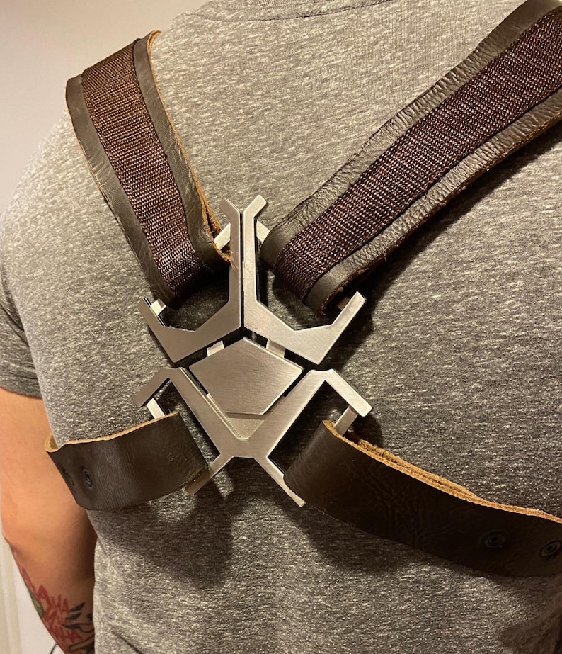 May include: A brown leather harness with a silver metal spider-shaped buckle. The harness has two straps that cross over the chest and attach to the buckle.