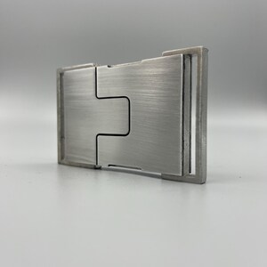 May include: A brushed metal money clip with a rectangular shape and a unique interlocking design. The clip has a silver finish and a minimalist aesthetic, suitable for holding cash and cards.