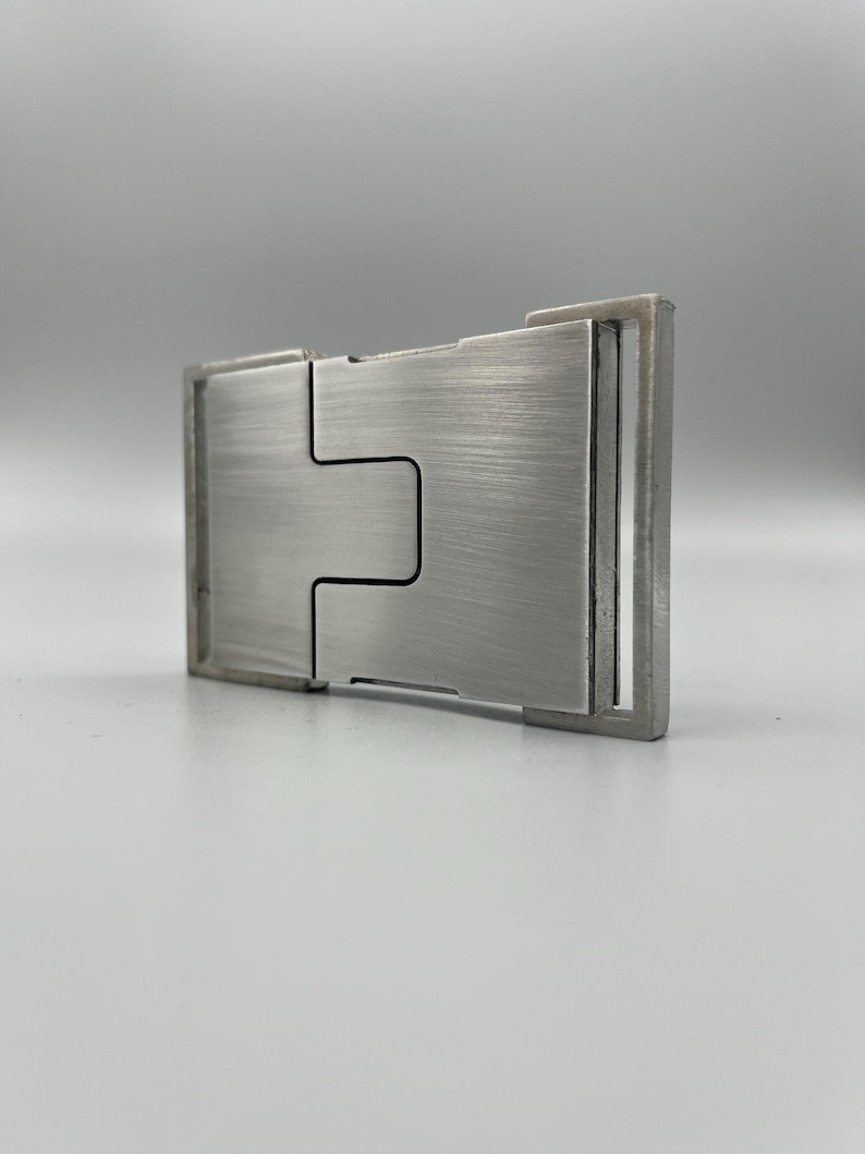May include: A brushed metal money clip with a rectangular shape and a unique interlocking design. The clip has a silver finish and a minimalist aesthetic, suitable for holding cash and cards.