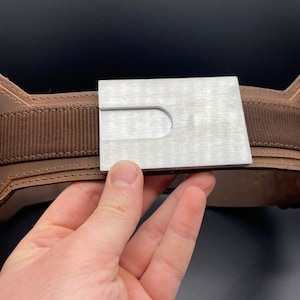 May include: A brown leather belt with a silver metal buckle. The buckle has a rectangular shape with a cut-out in the center.