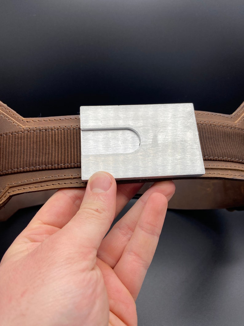 May include: A brown leather belt with a silver metal buckle. The buckle has a rectangular shape with a cut-out in the center.