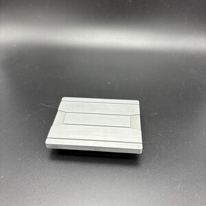 Pode incluir: Um objeto quadrado cinza claro com uma superf&iacute;cie texturizada e linhas horizontais. O objeto tem uma borda ligeiramente elevada e parece ser feito de um material s&oacute;lido. O fundo &eacute; de cor cinza escuro.