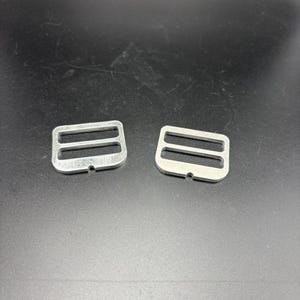 Puede incluir: Dos hebillas correderas met&aacute;licas plateadas, cada una con dos ranuras. Las hebillas son de metal brillante y est&aacute;n sobre un fondo oscuro. Estas hebillas se utilizan com&uacute;nmente en proyectos de costura y artesan&iacute;a.