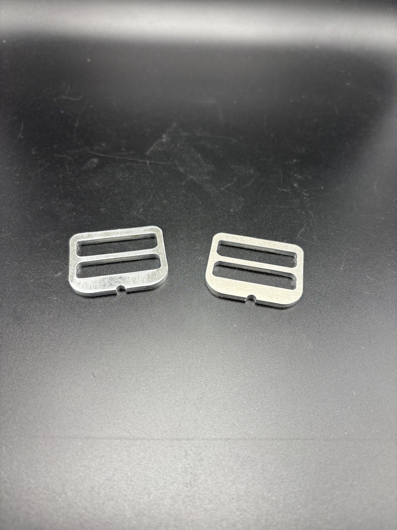 Puede incluir: Dos hebillas correderas met&aacute;licas plateadas, cada una con dos ranuras. Las hebillas son de metal brillante y est&aacute;n sobre un fondo oscuro. Estas hebillas se utilizan com&uacute;nmente en proyectos de costura y artesan&iacute;a.