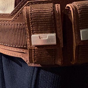 Op de afbeelding: Close-up van een bruine leren riem met twee zakjes en zilverkleurige metalen accenten. De riem heeft een getextureerd, geribbeld ontwerp en is gestikt met bruin garen. De achtergrond toont een marineblauwe stof.