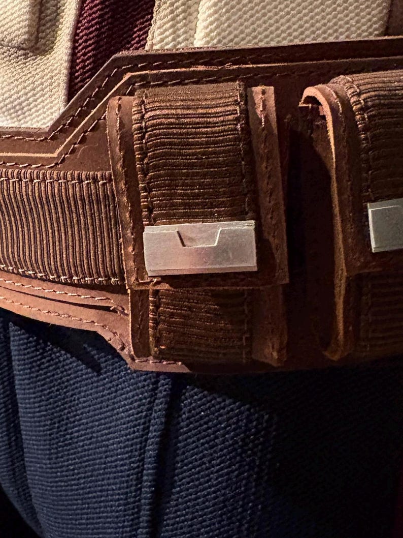 Op de afbeelding: Close-up van een bruine leren riem met twee zakjes en zilverkleurige metalen accenten. De riem heeft een getextureerd, geribbeld ontwerp en is gestikt met bruin garen. De achtergrond toont een marineblauwe stof.
