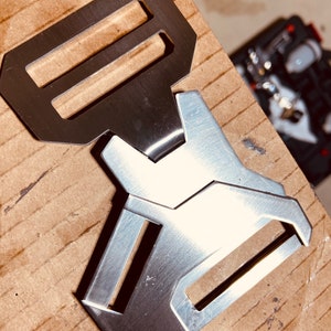 May include: A silver metal buckle with two slots. The buckle is designed with a geometric shape and has a brushed finish.