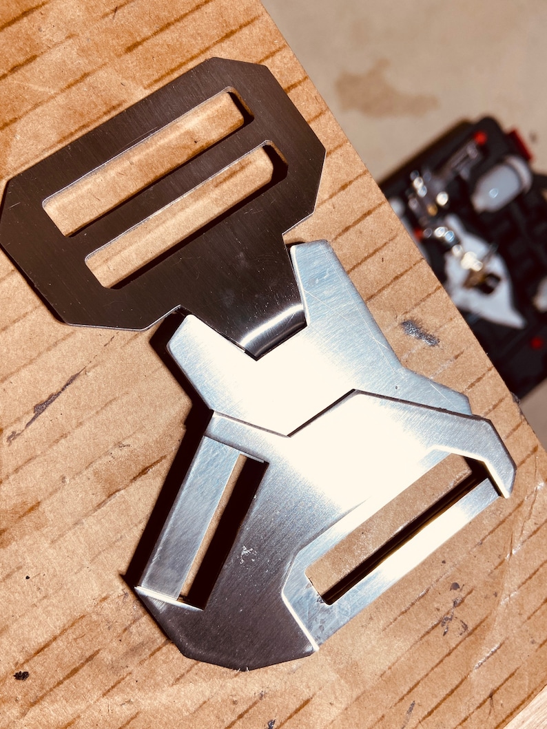 May include: A silver metal buckle with two slots. The buckle is designed with a geometric shape and has a brushed finish.