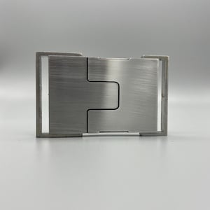 May include: A brushed metal belt buckle with a rectangular frame and a central, interlocking design. The buckle has a matte finish and a minimalist aesthetic, suitable for various belt styles.