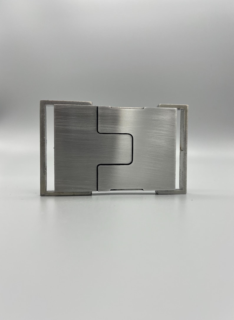May include: A brushed metal belt buckle with a rectangular frame and a central, interlocking design. The buckle has a matte finish and a minimalist aesthetic, suitable for various belt styles.
