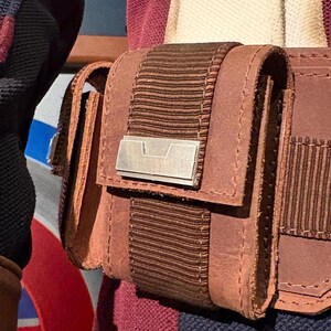 Op de afbeelding: Close-up van een bruine leren tas met een klep en een zilverkleurig metalen accent. De tas is bevestigd aan een bruine riem met een geribbeld stoffen detail. De stiksels zijn zichtbaar en het ontwerp suggereert een vintage of tactische esthetiek.