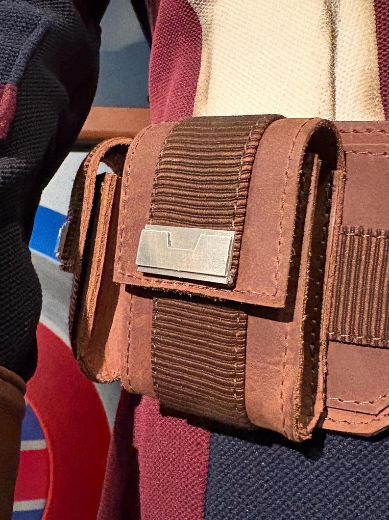Op de afbeelding: Close-up van een bruine leren tas met een klep en een zilverkleurig metalen accent. De tas is bevestigd aan een bruine riem met een geribbeld stoffen detail. De stiksels zijn zichtbaar en het ontwerp suggereert een vintage of tactische esthetiek.