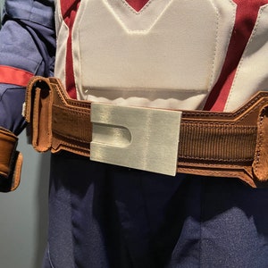 May include: A brown leather belt with a silver buckle. The belt has a textured fabric and a design on the buckle.