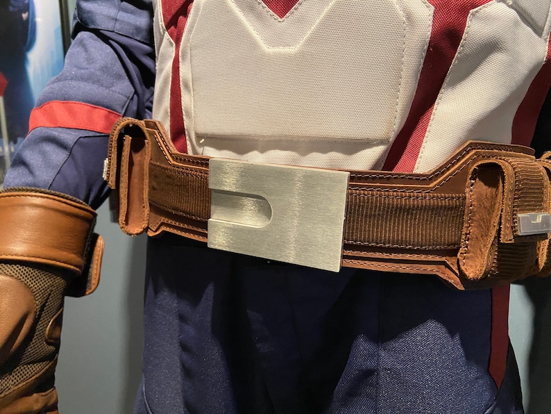 May include: A brown leather belt with a silver buckle. The belt has a textured fabric and a design on the buckle.
