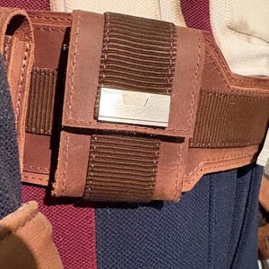 Op de afbeelding: Close-up van een bruine leren riem met een klein zakje en een zilverkleurig metalen accent. Het zakje heeft een geribbeld bruin stoffen detail. De riem wordt gedragen over een marineblauw kledingstuk en een bordeauxrode stof.