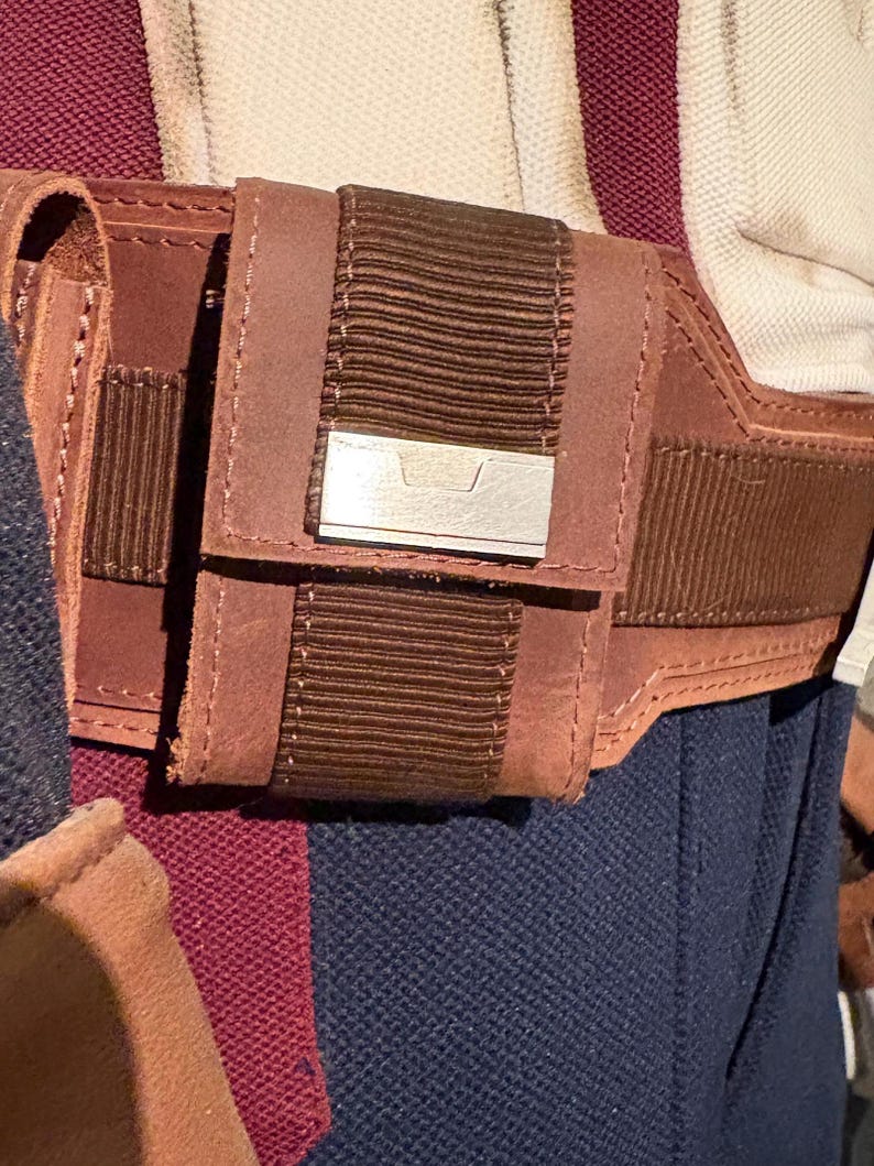 Op de afbeelding: Close-up van een bruine leren riem met een klein zakje en een zilverkleurig metalen accent. Het zakje heeft een geribbeld bruin stoffen detail. De riem wordt gedragen over een marineblauw kledingstuk en een bordeauxrode stof.