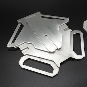 May include: A silver metal phone stand with a geometric design. The stand has a slot for holding a phone upright.