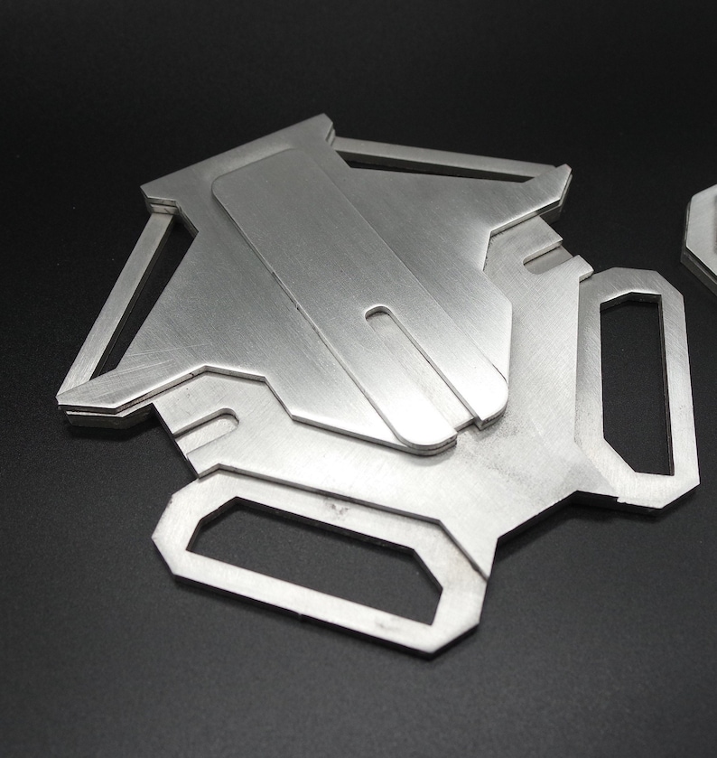 May include: A silver metal phone stand with a geometric design. The stand has a slot for holding a phone upright.