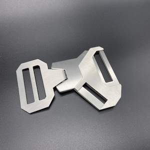 May include: A silver metal buckle with a rectangular shape and two slots. The buckle is designed to be used with a strap or belt.