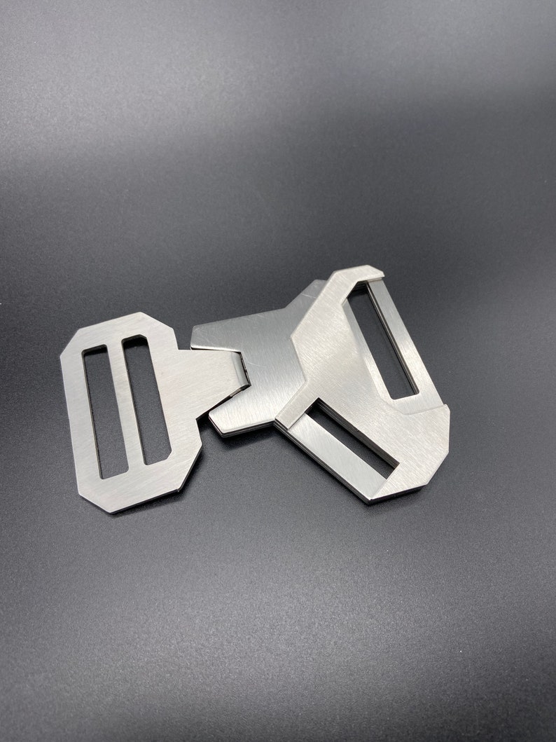 May include: A silver metal buckle with a rectangular shape and two slots. The buckle is designed to be used with a strap or belt.
