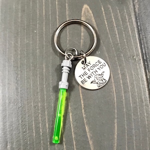 May include: A silver keyring with a green lightsaber charm and a round silver charm with the text "May the Force be with you" and a Yoda image.