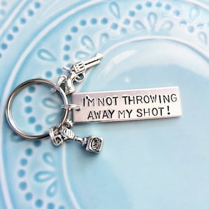 Könnte beinhalten: Silberfarbener Schlüsselanhänger mit einem Revolver-Anhänger, einem kleinen Tintenflaschen-Anhänger und einem rechteckigen Anhänger mit der Aufschrift "I'M NOT THROWING AWAY MY SHOT!". Der Schlüsselanhänger liegt auf einer hellblauen Oberfläche.