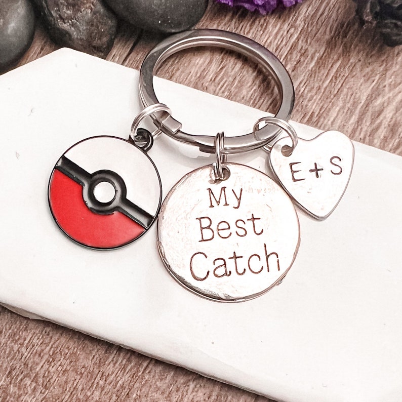 May include: A silver keychain with three charms: a red and white Pokeball, a silver charm with the text "My Best Catch", and a heart-shaped charm with the text "E+S".