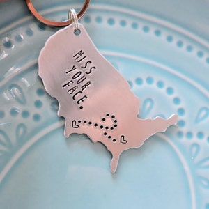 Puede incluir: Un llavero de plata con forma de Estados Unidos con las palabras "Miss Your Face" y una línea punteada con corazones en cada extremo.