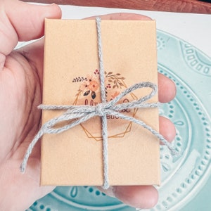 Pu&ograve; includere: Una piccola scatola marrone legata con un nastro grigio e un logo floreale che dice "Little Things Boutique".
