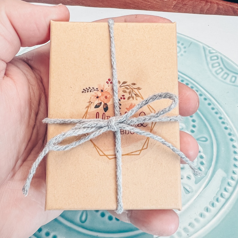 Pu&ograve; includere: Una piccola scatola marrone legata con un nastro grigio e un logo floreale che dice "Little Things Boutique".