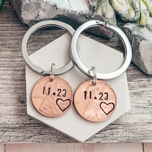 May include: Two silver keychains with copper penny charms. Each penny has the date 11.23 and a heart with the year 2018 on one and 2013 on the other.