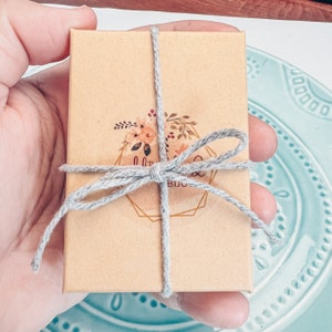 May include: A small, brown cardboard box tied with a gray ribbon and a bow. The box has a floral design with the text "Little Things Boutique" printed on it.