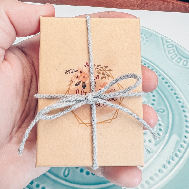 May include: A small, brown cardboard box tied with a gray ribbon and a bow. The box has a floral design with the text "Little Things Boutique" printed on it.