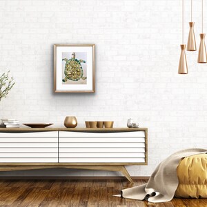 K&ouml;nnte beinhalten: Ein gerahmtes Aquarellbild einer gr&uuml;nen und goldenen Schildkr&ouml;te auf wei&szlig;em Hintergrund. Das Bild h&auml;ngt an einer wei&szlig;en Backsteinwand &uuml;ber einer wei&szlig;en Kommode mit goldenen Akzenten.