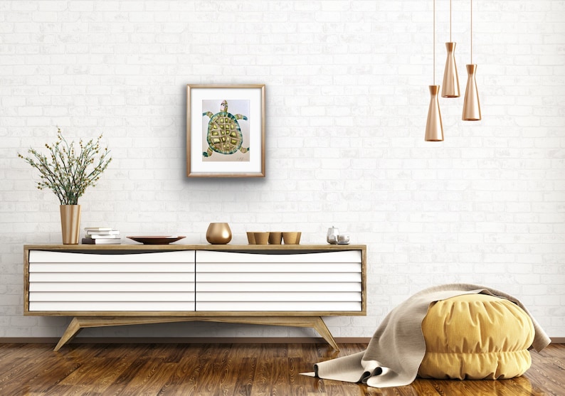 K&ouml;nnte beinhalten: Ein gerahmtes Aquarellbild einer gr&uuml;nen und goldenen Schildkr&ouml;te auf wei&szlig;em Hintergrund. Das Bild h&auml;ngt an einer wei&szlig;en Backsteinwand &uuml;ber einer wei&szlig;en Kommode mit goldenen Akzenten.