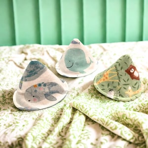 May include: Three cone-shaped fabric covers featuring printed illustrations of a blue whale, a grey narwhal, and a green jungle scene with a giraffe and a brown bear. The items are constructed from soft textile material with stitched edges.