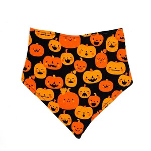 May include: A triangular bandana featuring a black background with a repeating pattern of orange pumpkins. Each pumpkin displays a unique, simple facial expression with black eyes and mouths, creating a festive design across the fabric surface.