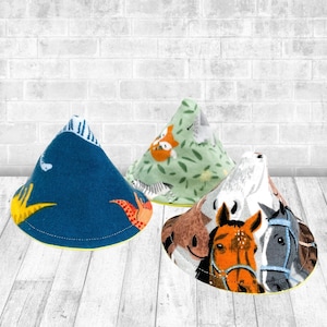 May include: Three cone-shaped fabric covers featuring distinct patterns: a dark blue dinosaur print, a light green jungle animal print with tigers and zebras, and a white, brown, and black horse print. Each item has a circular base and pointed top.