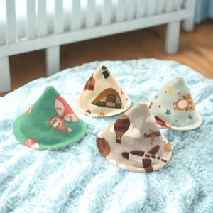 May include: Four cone-shaped fabric covers featuring various prints: a green cover with a fox, a cream cover with a tent, a gray cover with airplanes and hot air balloons, and a light blue cover with a bear, moon, and stars pattern.