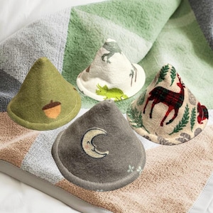 May include: Four cone-shaped fabric items with various designs, including an acorn, dinosaurs, a deer, and a crescent moon. The items are displayed on a patterned blanket. Text at the bottom reads "Over 20 designs to choose from."