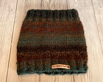 May include: A hand-knitted dog cowl in shades of brown, teal, and rust. The cowl features a ribbed texture and a small rectangular tag that reads "The Highland Hound". The item is made of a soft, textured yarn.