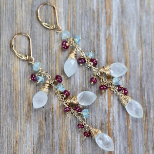 May include: Gold earrings with a dangling design featuring white and blue gemstones. The earrings are made with wire wrapped around the gemstones and have a delicate, bohemian style.