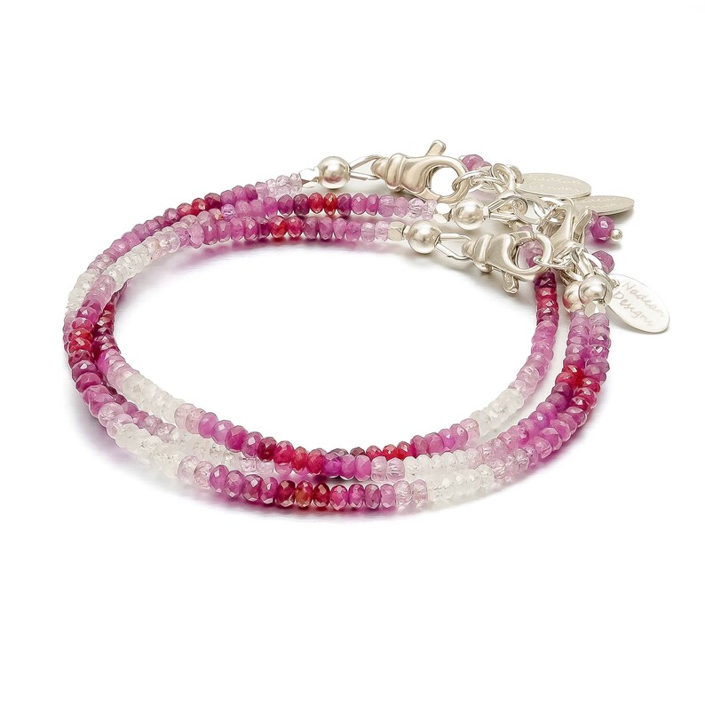May include: A delicate multi-strand bracelet featuring a gradient of pink, red, and white gemstones. The bracelet is made with small, round beads and has a silver clasp and a small tag that reads "Nadine Design".