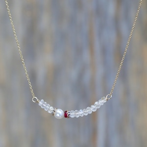 May include: A delicate gold chain necklace featuring a curved bar pendant. The pendant is adorned with a central white pearl, a small red gemstone, and clear, faceted beads. The necklace is a simple, elegant piece of jewelry.