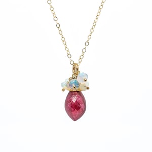 May include: A gold chain necklace with a pendant featuring a large, faceted, red ruby gemstone surrounded by smaller, white opal gemstones.