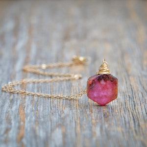 May include: A gold chain necklace with a pink gemstone pendant wrapped in gold wire. The pendant is a rough, natural-looking stone.