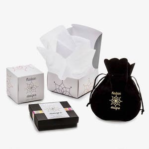 May include: Three boxes with the text "Nadean Designs" on them. The boxes are white with a colorful geometric pattern. One box is open and filled with white tissue paper. The other two boxes are closed. There is also a black velvet pouch with the text "Nadean Designs" on it.