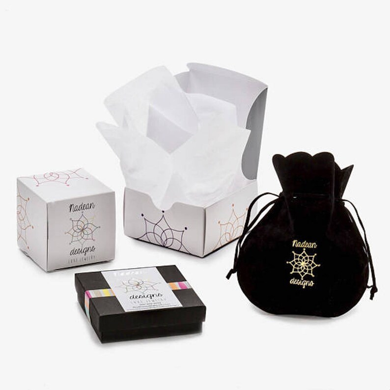May include: Three boxes with the text "Nadean Designs" on them. The boxes are white with a colorful geometric pattern. One box is open and filled with white tissue paper. The other two boxes are closed. There is also a black velvet pouch with the text "Nadean Designs" on it.